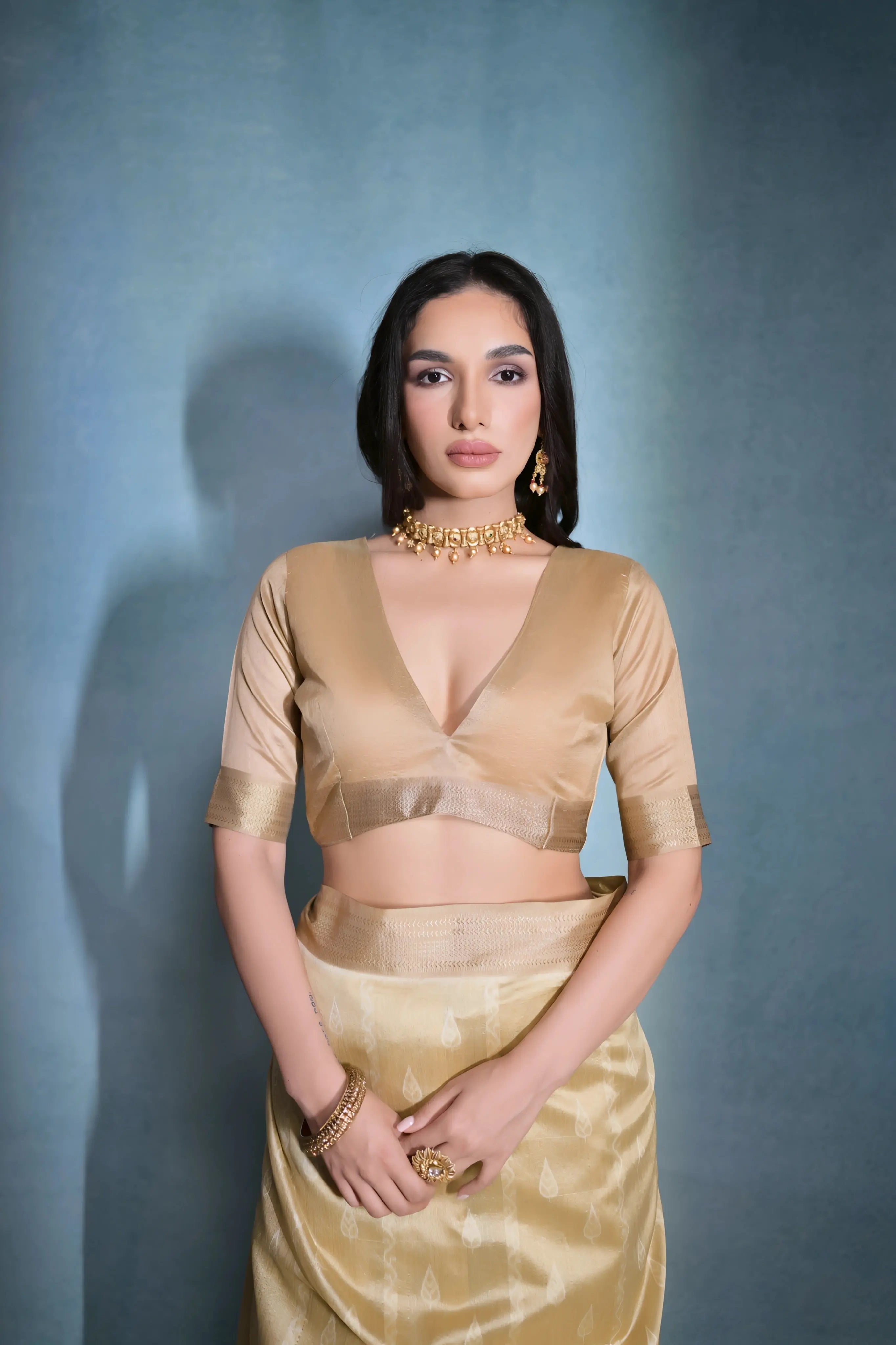 A woman models an elegant Beige/Champagne Soft Raw Silk Saree with a wide metallic gold Zari border. The pallu features distinct horizontal bands of dark brown/black Ikat-style geometric weaving, providing a strong contrast. She wears a matching beige blouse and accessorizes with a gold choker necklace and large gold earrings, posing indoors.